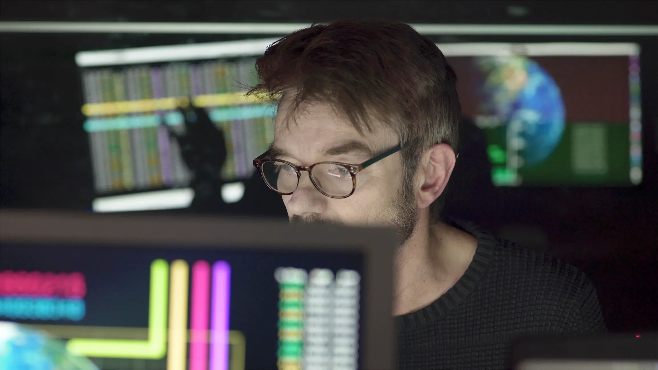 Man with glasses focused on computer screens displaying colorful data, charts, and a globe in a dark room.