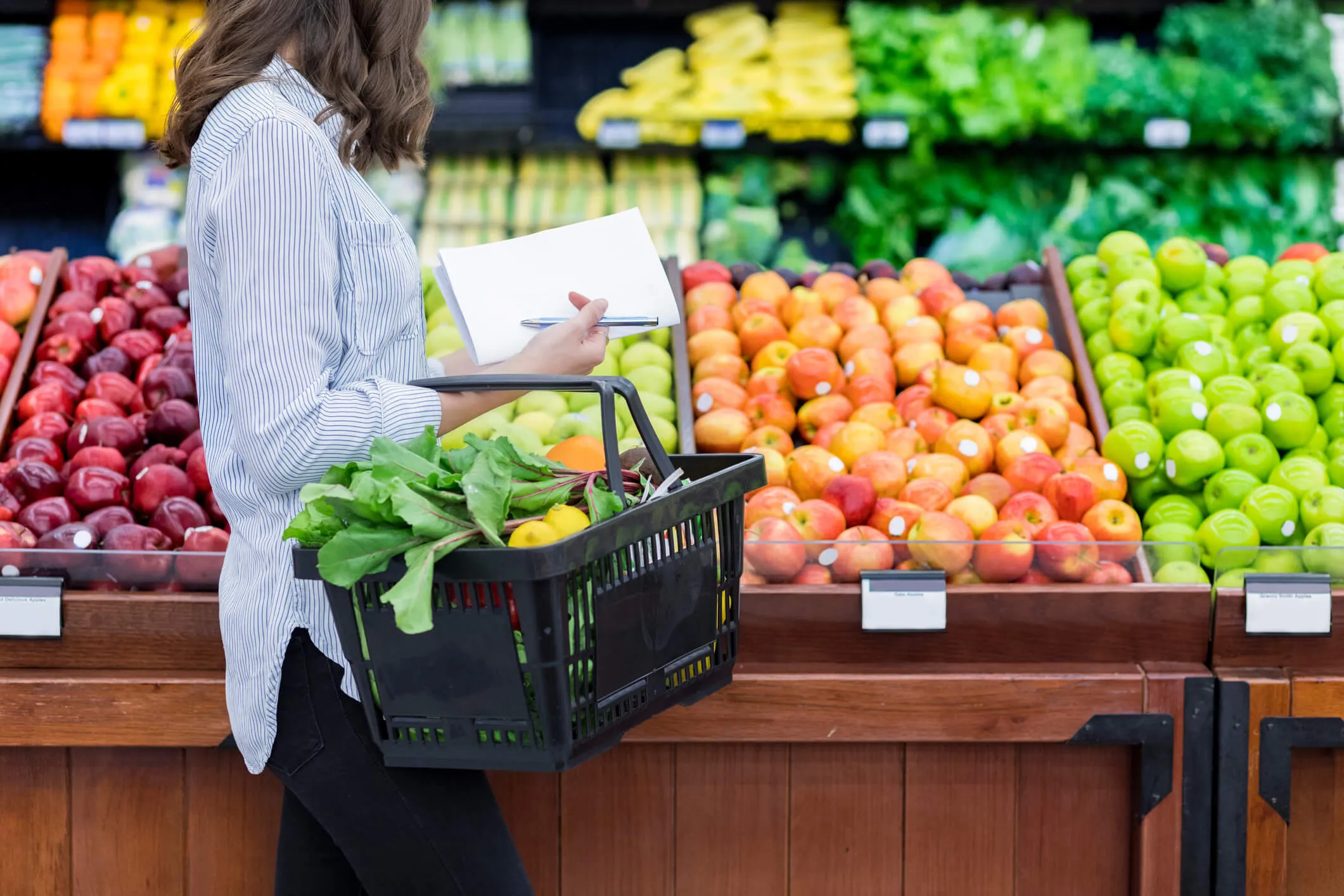 Person holding a shopping basket filled with fresh vegetables and fruit while checking a shopping list in a produce aisle with apples, bananas, and leafy greens.