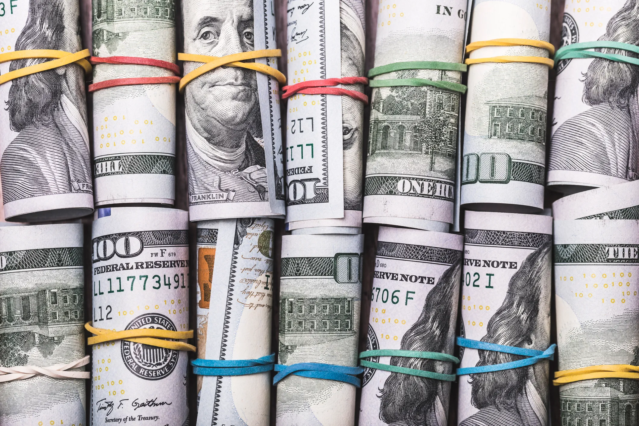 Rolled U.S. hundred-dollar bills secured with rubber bands of various colors arranged closely together.
