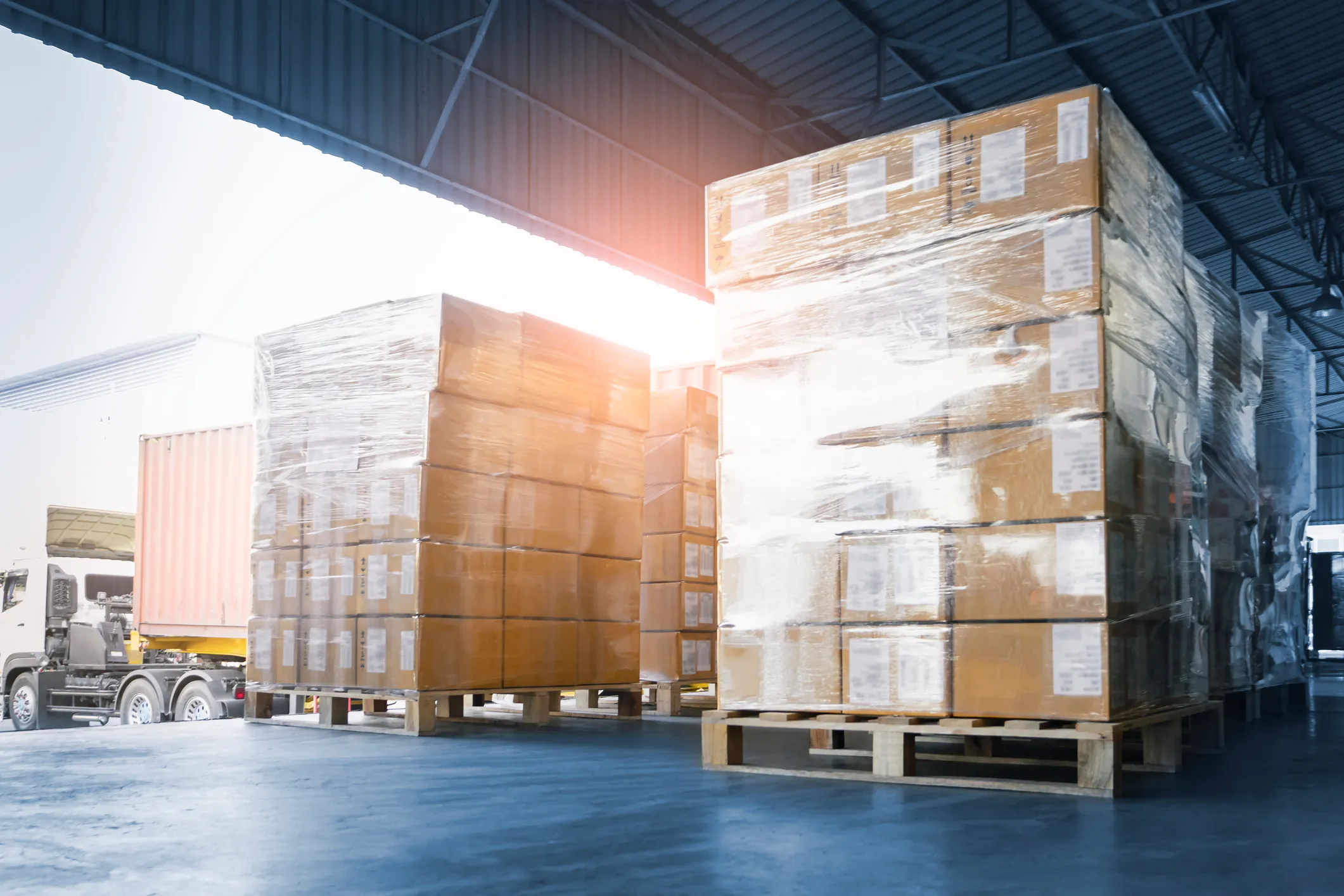 Large stacks of cardboard boxes shrink-wrapped on wooden pallets inside a warehouse near a semi-truck with a shipping container attached.