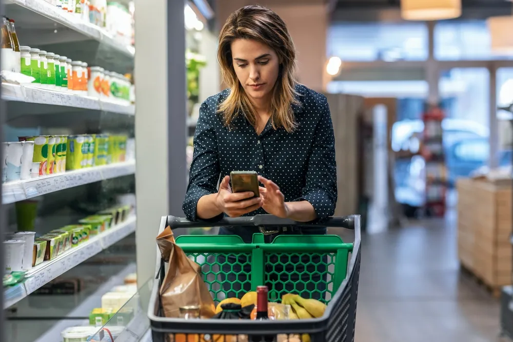 Woman with shoulder-length hair wearing a dark blue polka dot shirt pushes a shopping cart with groceries including bananas and a bottle, while looking at her smartphone in a grocery store aisle.