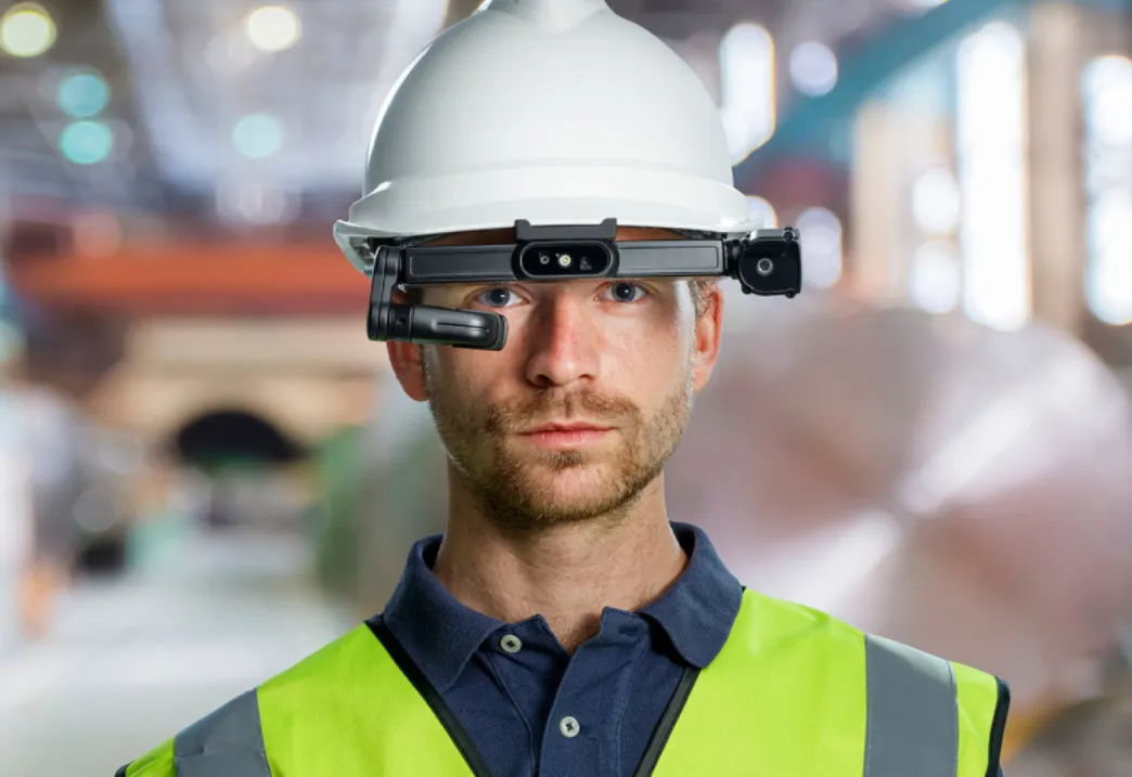 Man wearing a white hard hat and yellow safety vest with a smart glasses headset in an industrial warehouse setting.
