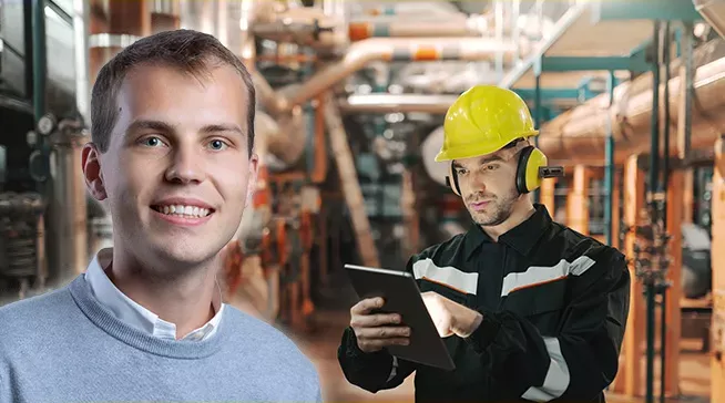A man in a gray sweater smiling at the camera beside a worker in a yellow hard hat and protective earmuffs using a tablet inside an industrial facility with pipes.