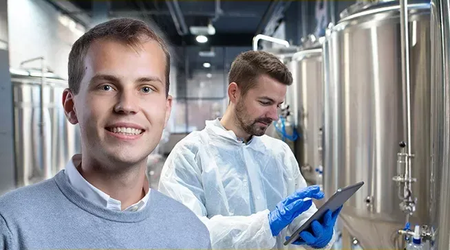 Two men in a brewery or beverage production facility with large stainless steel tanks; one man in a gray sweater smiles at the camera, while the other wears a white protective suit and blue gloves, interacting with a tablet.