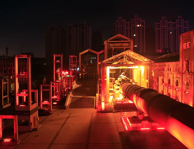 Industrial scene at night featuring a large, illuminated rotary kiln or cylindrical machinery with bright orange-red lighting, surrounded by buildings and tall high-rise structures in the background under a dark sky.