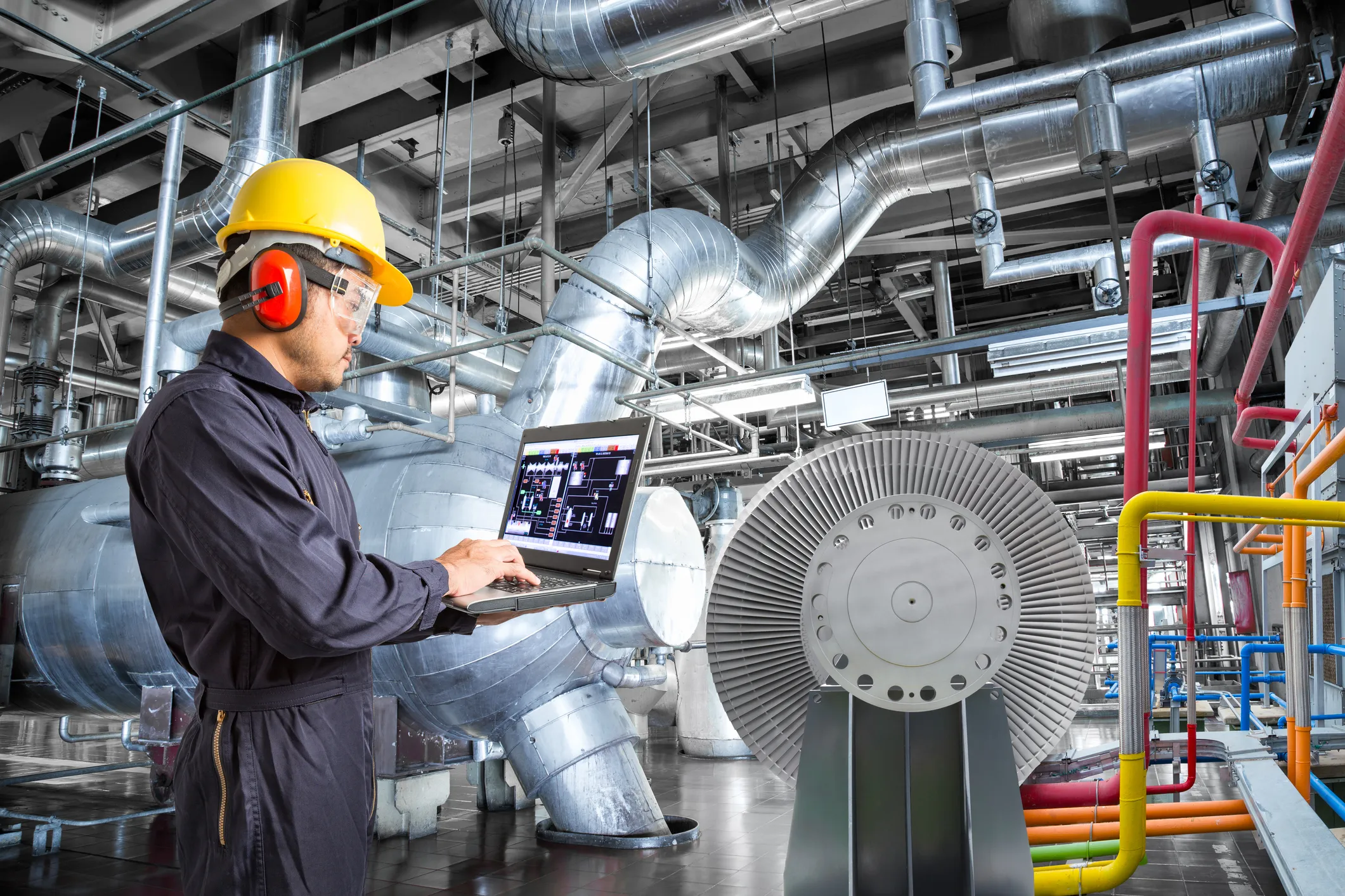 Industrial engineer wearing a yellow hard hat, safety goggles, and red ear protection using a laptop in a factory with large metal pipes, ducts, and machinery.