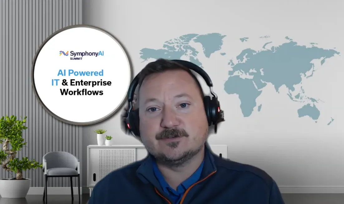 A man wearing a headset and a blue jacket in a modern office setting with a world map on the wall and a round sign displaying 'SymphonyAI SUMMIT AI Powered IT & Enterprise Workflows'.