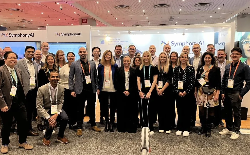 Group photo of 24 diverse SymphonyAI team members standing together at an exhibit booth with SymphonyAI banners and screens in the background, all wearing conference badges.
