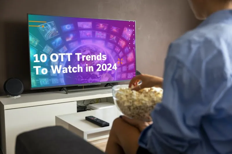 Person sitting on a couch holding a bowl of popcorn, watching a TV screen displaying the text '10 OTT Trends To Watch in 2024' with colorful digital backgrounds of screens and data.