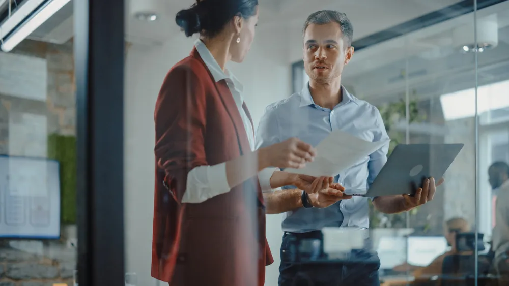 A man holding a laptop and a woman holding a paper are having a discussion in a modern office with glass walls.