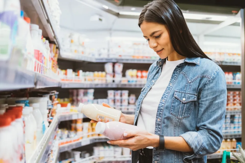 Woman in a denim jacket compares two bottles of dairy or plant-based drinks while standing in a grocery store aisle with shelves stocked with various beverages.