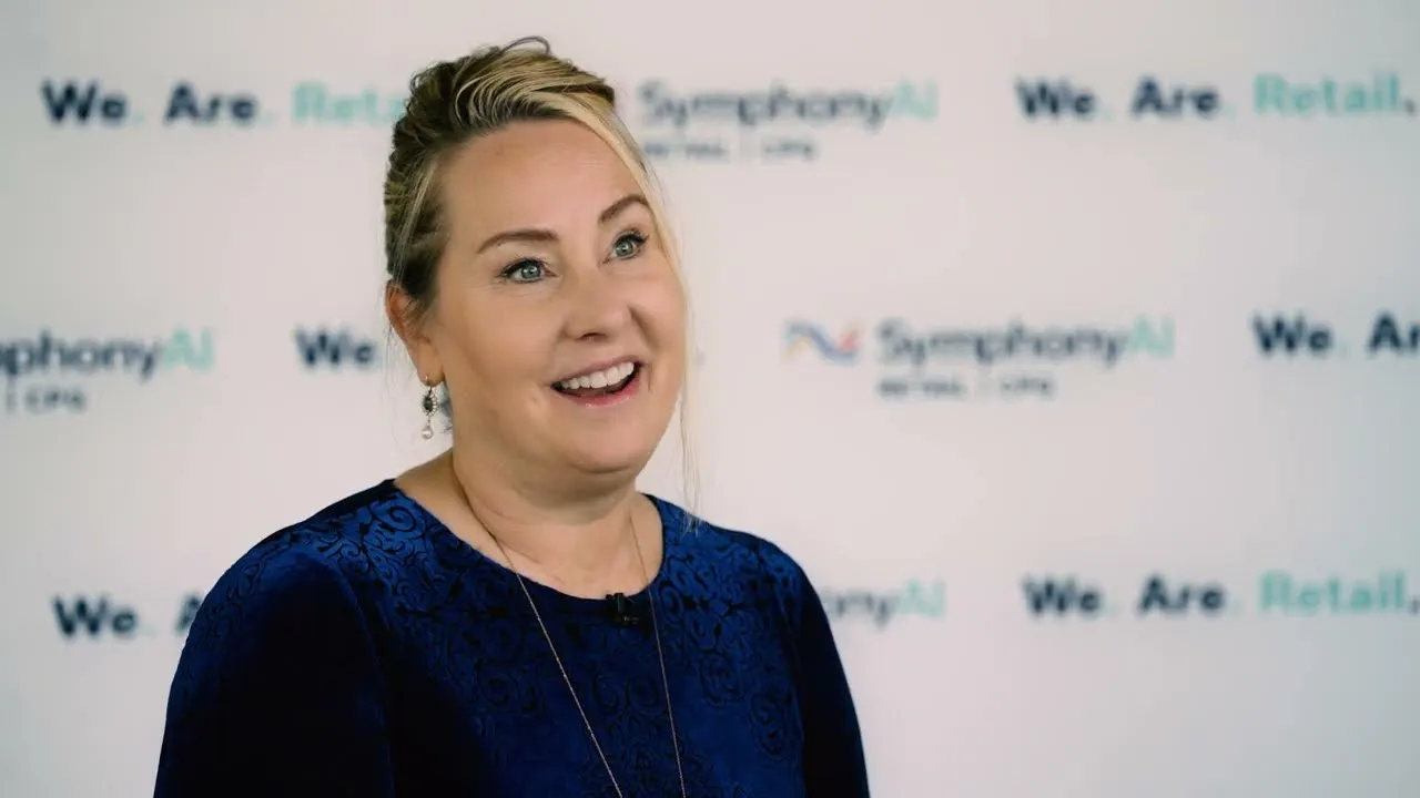 Smiling woman with blonde hair pulled back wearing a dark blue patterned top, standing in front of a white backdrop with repeated logos and text reading 'We Are Retail, SymphonyAI'.