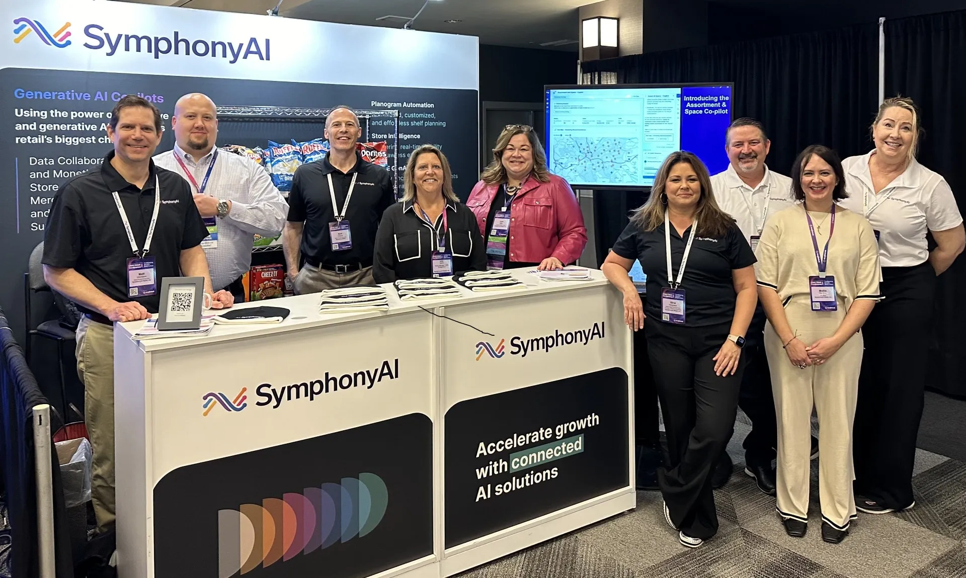 Nine people standing behind and beside a SymphonyAI booth at a professional event, smiling and wearing event badges and SymphonyAI-branded shirts, with promotional materials on the counter and a screen showing product information in the background.