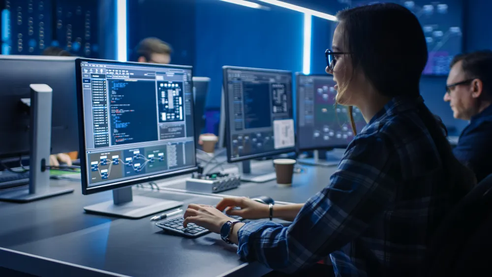 Woman wearing glasses and a plaid shirt typing on a keyboard while working on computer monitors displaying software code and graphical interfaces in a modern office with colleagues in the background.