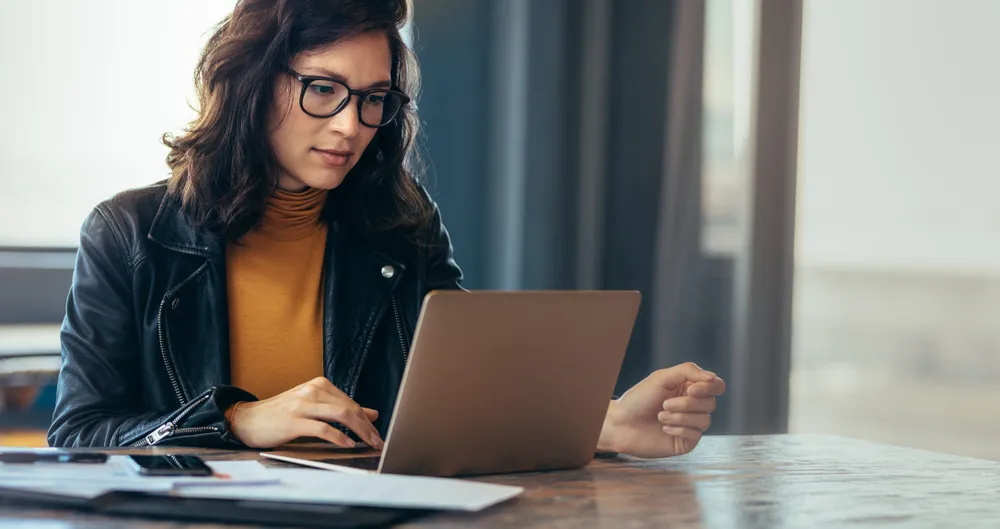 A woman with dark hair and glasses wearing a black leather jacket and mustard turtleneck is focused on her laptop at a wooden table indoors, with papers and a smartphone nearby.