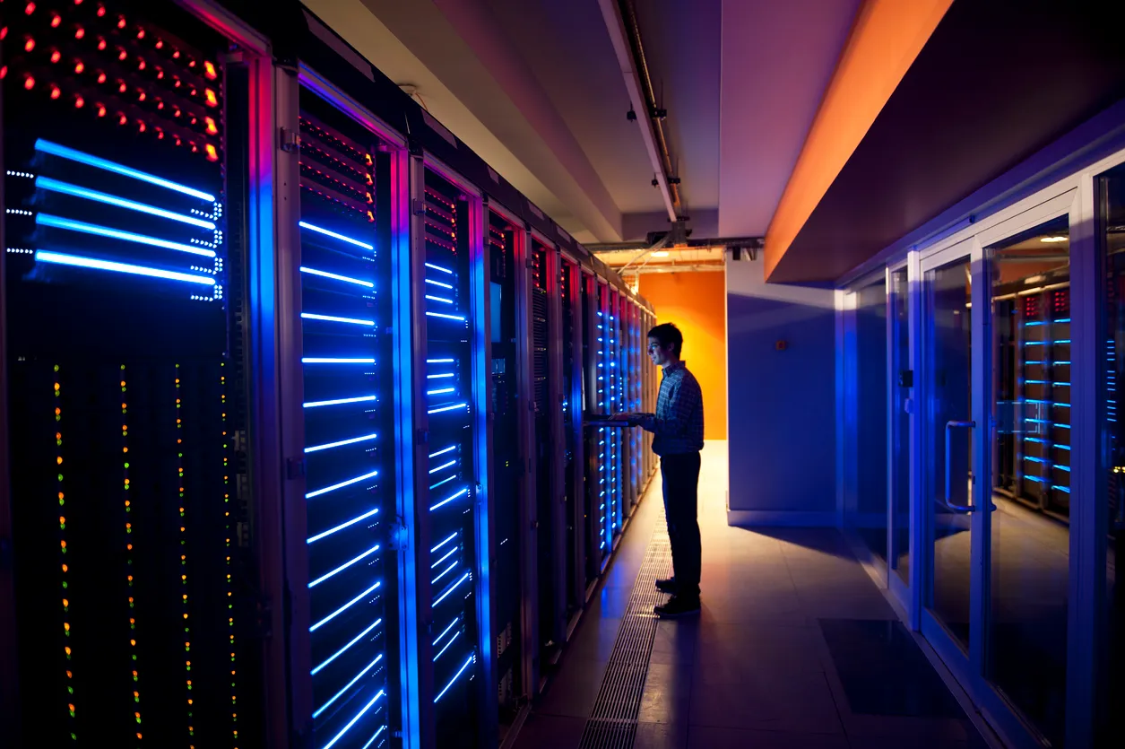 Person standing and working on a laptop in a server room illuminated by blue and red LED lights on server racks.