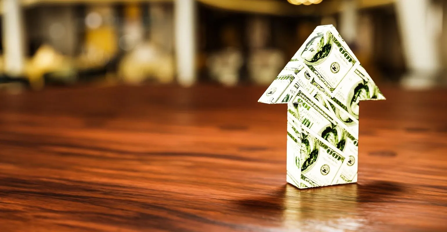 Arrow pointing upward made from folded hundred-dollar bills standing on a wooden surface with a blurred background, symbolizing financial growth.