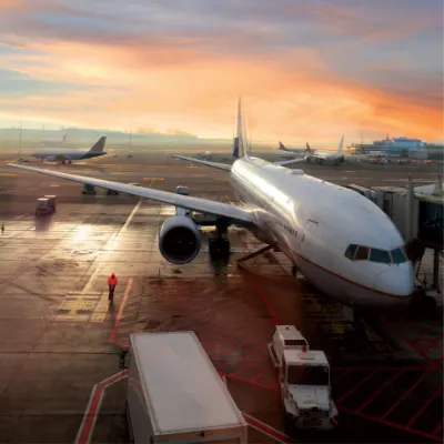 A large airplane at an airport gate during sunset with ground vehicles nearby and other planes in the background on the runway.