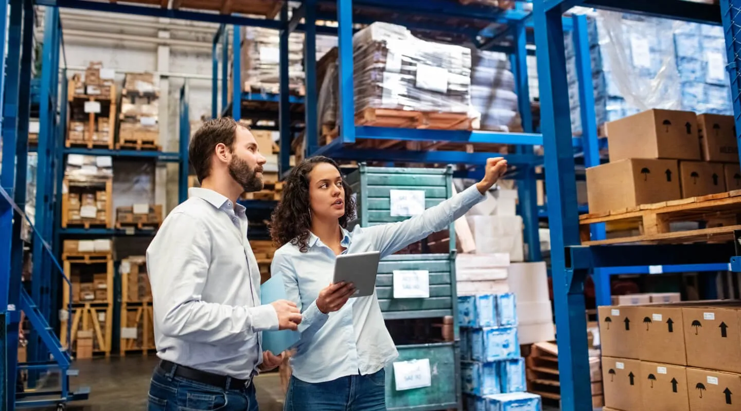 Two warehouse workers, a man and woman in light blue shirts, discussing while the woman points towards stacked shelves filled with boxes and pallets, the man holding a folder and the woman holding a tablet.