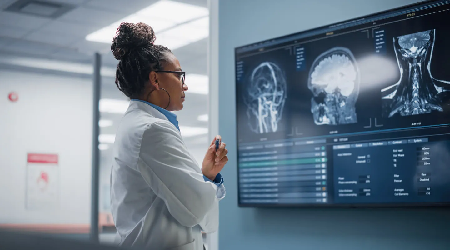Female doctor in a white coat analyzing brain and neck MRI scans displayed on a large digital screen in a medical facility.