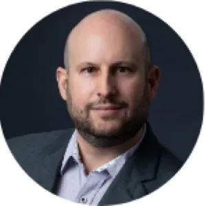 Portrait of a bald man with a trimmed beard wearing a gray suit jacket and light-colored shirt against a dark background.