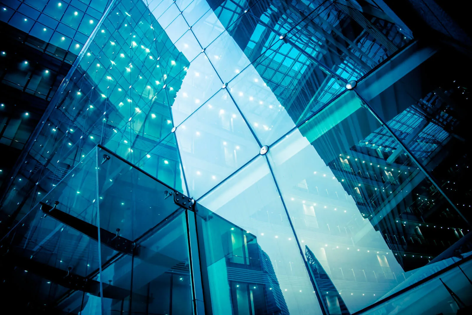 Close-up view of a modern glass building facade with reflections of structural elements and illuminated lights creating a blue-toned abstract pattern.