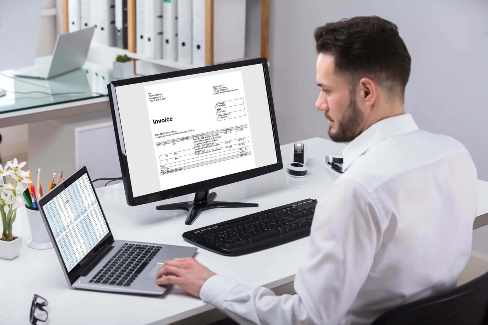Man in white shirt sitting at desk working on laptop and desktop computer showing invoice and spreadsheet data, with office supplies and plants on the desk.