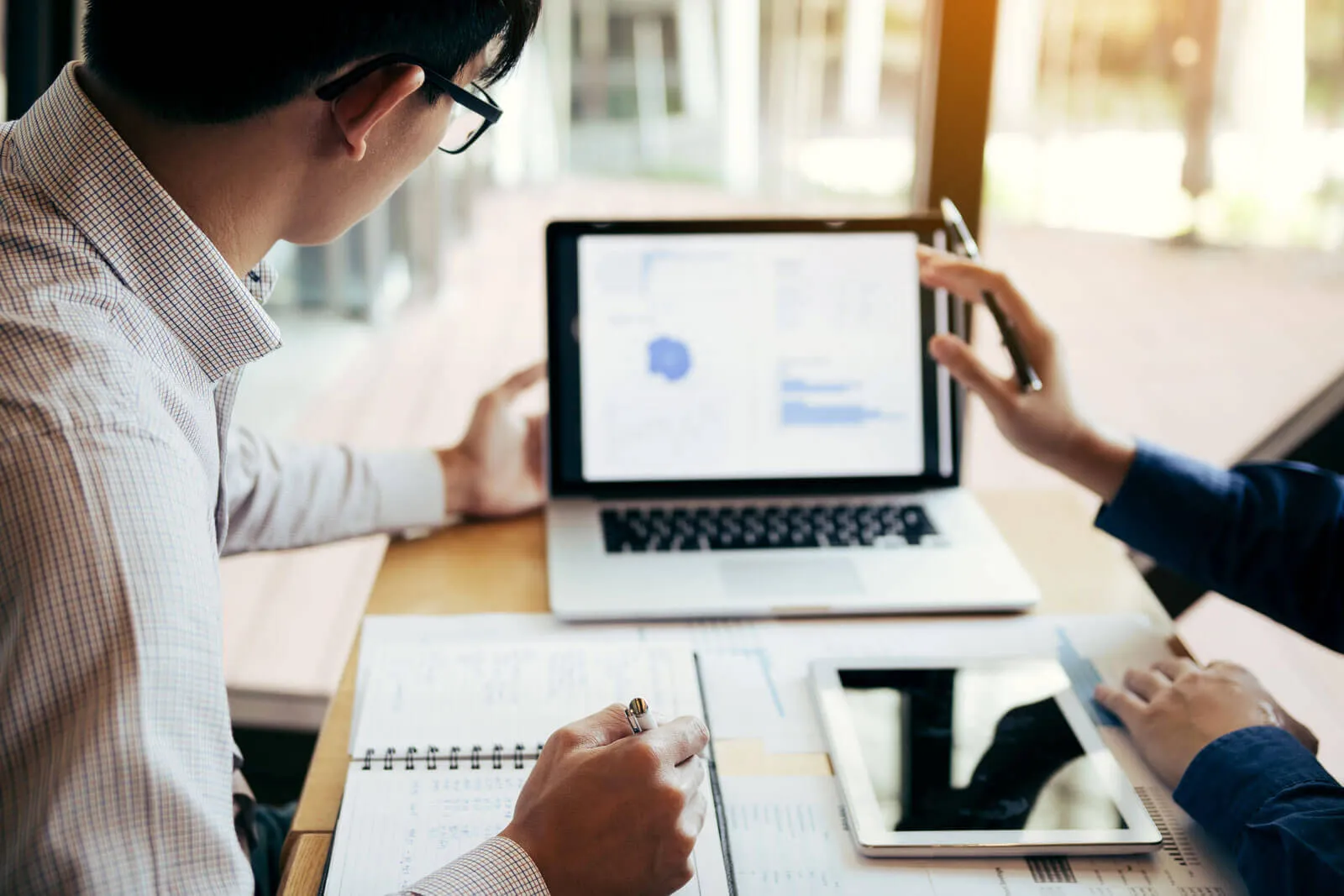 Two people working at a desk with documents, a tablet, and a laptop showing blurred charts and data, one holding a pen and the other pointing at the laptop screen.