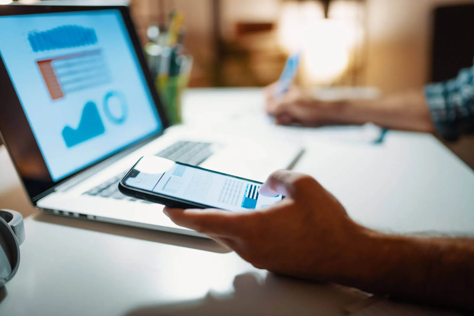 Person holding a smartphone displaying charts and graphs in front of a laptop with blurred data visuals on the screen, while writing on paper in the background.