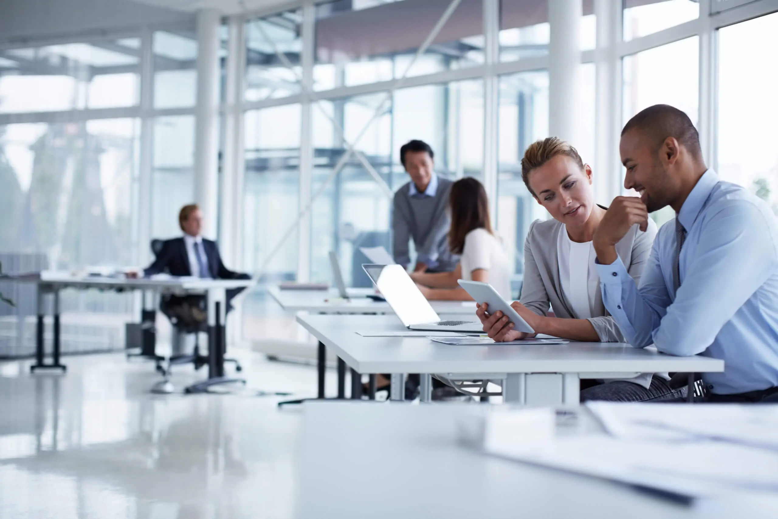 Two business colleagues sitting and discussing while looking at a tablet in a modern, bright office with other people working in the background.