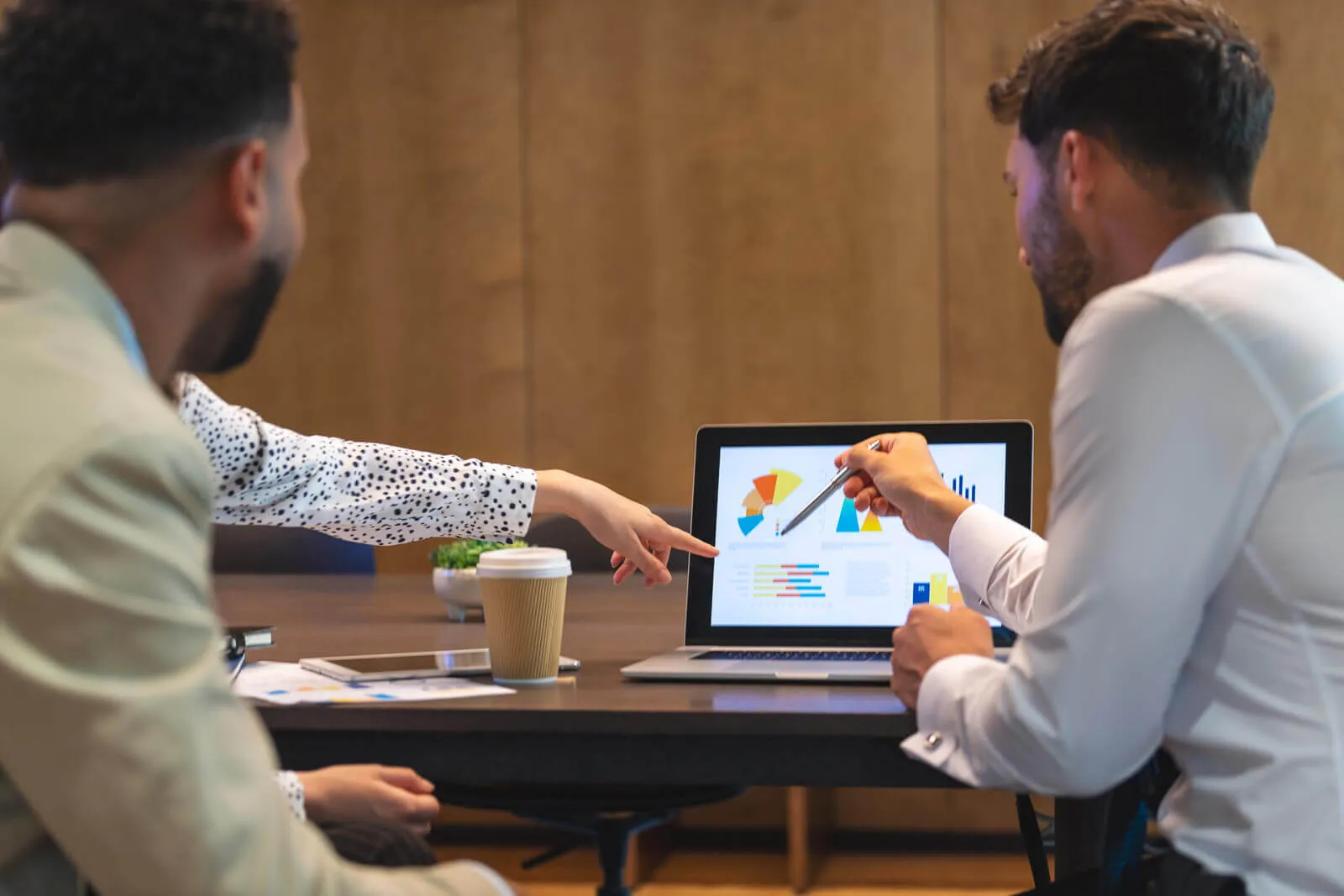 Three professionals in a meeting pointing at a laptop screen displaying colorful charts and graphs.