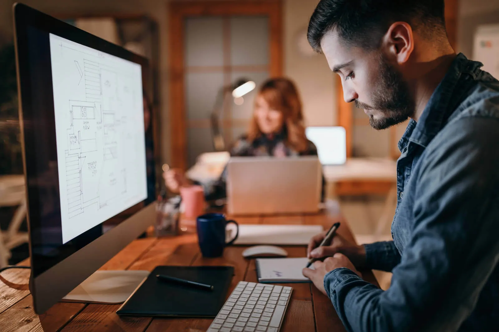 Man sitting at a wooden desk, writing notes on a pad beside a computer monitor displaying architectural floor plans, with a woman working on a laptop blurred in the background.