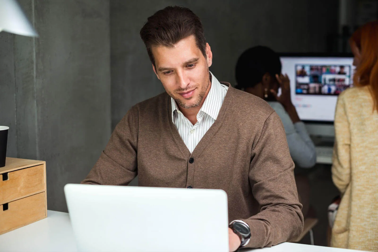 A man wearing a brown cardigan and white striped shirt is focused on working on a laptop at a desk, with two colleagues working on a desktop computer in the blurred background.