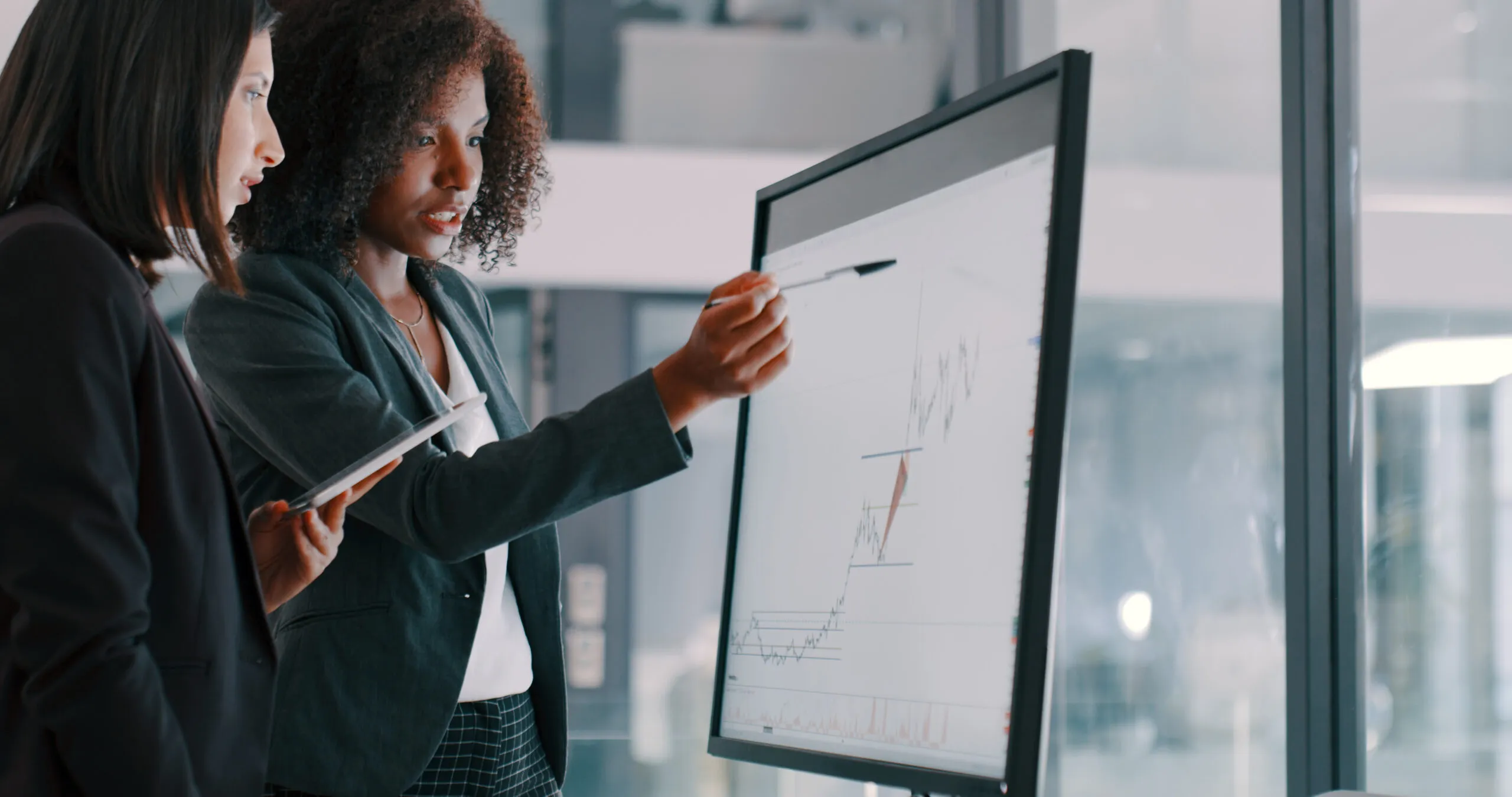 Two professional women discuss data on a large screen; one points at a rising graph with a pen while the other holds a tablet.