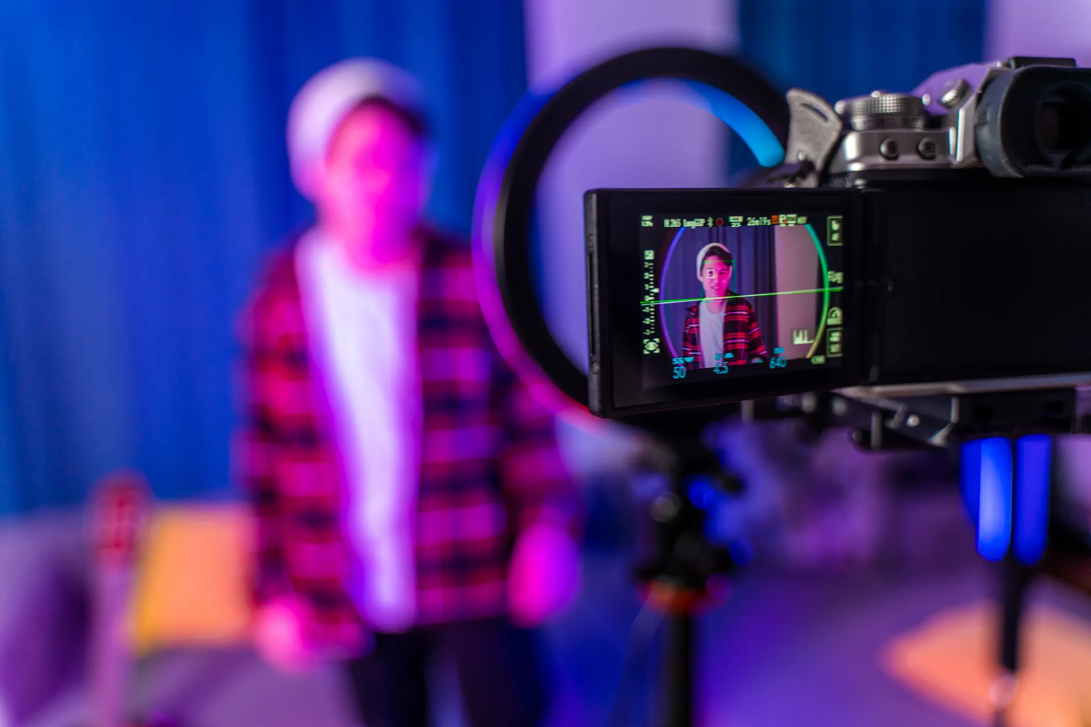 Camera screen showing a young person in a red checkered shirt and white beanie smiling while being recorded indoors under pink and blue lighting.