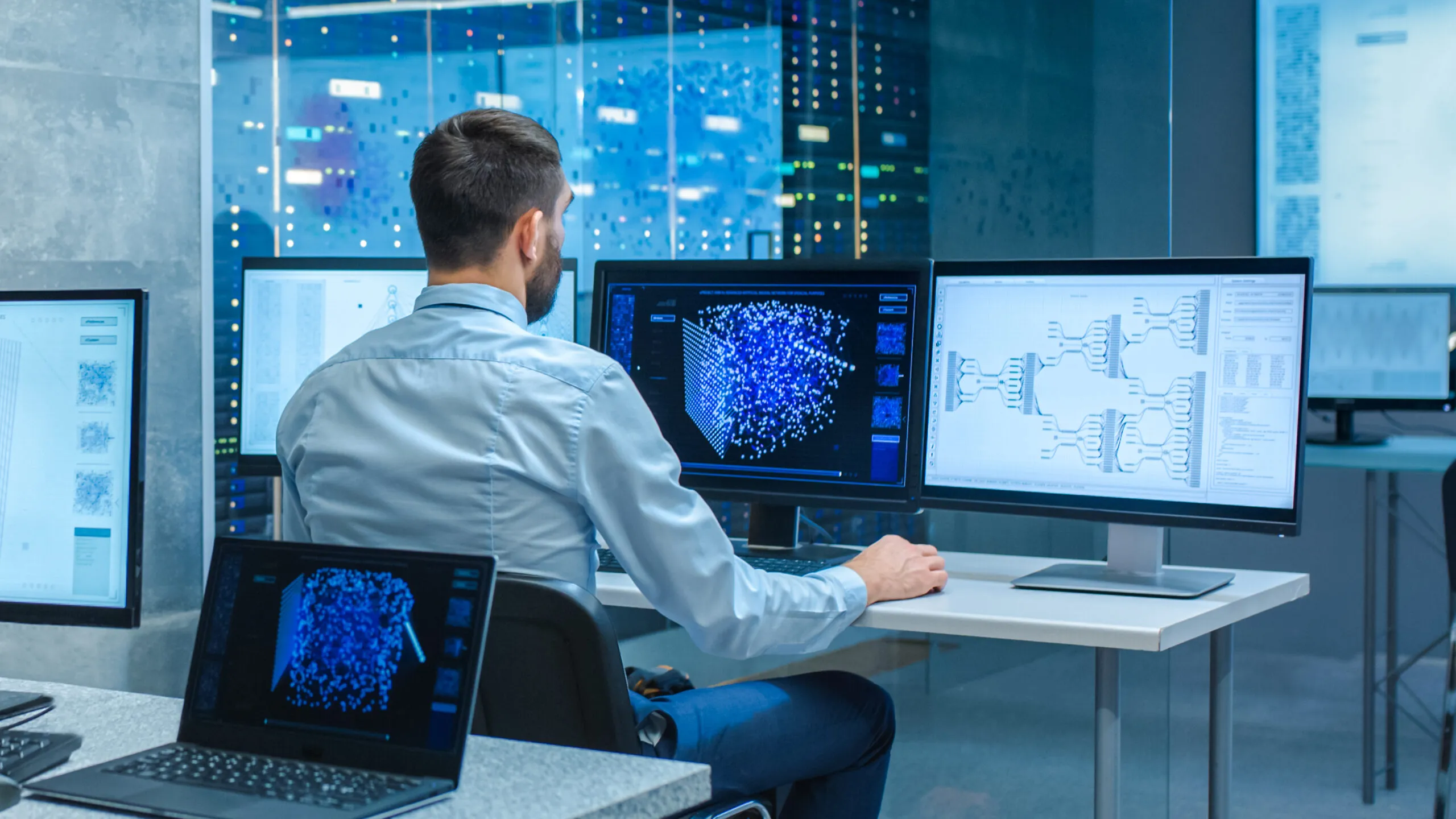 Man in a light blue shirt sitting at a desk working on a computer with three monitors displaying blue data visualizations and diagrams in a modern data center or tech office.
