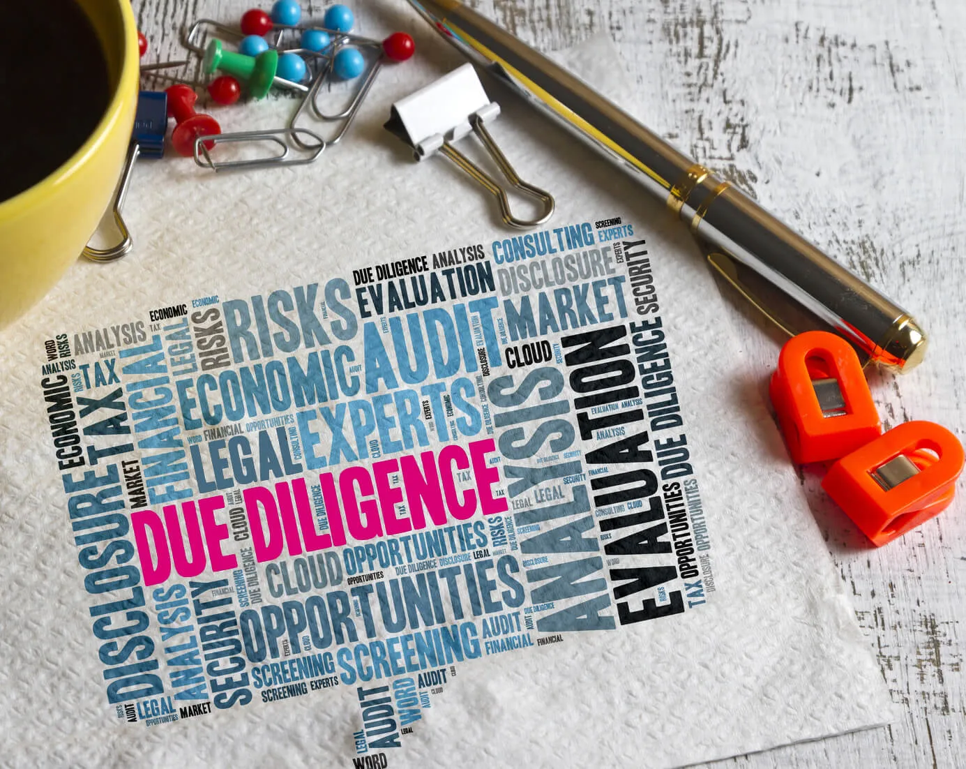 Word cloud with terms related to due diligence like 'Due Diligence', 'Audit', 'Evaluation', 'Analysis', 'Risks', and 'Opportunities' on a white paper towel, surrounded by a yellow coffee cup, colorful push pins, and a pen with two orange blade sharpeners on a wooden surface.