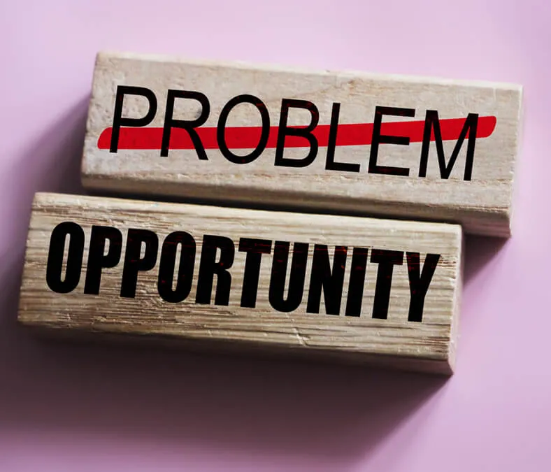 Two wooden blocks on a pink surface; the top block shows the word 'PROBLEM' with a red line crossing it out, and the bottom block shows the word 'OPPORTUNITY' in bold black letters.