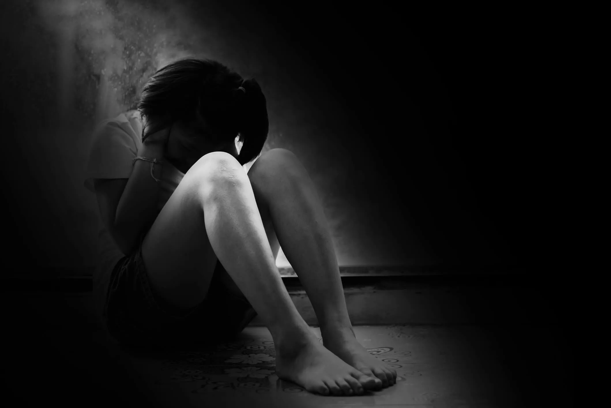 Black and white image of a young person sitting on the floor with knees drawn up and head bowed, appearing distressed or fearful in a dark, empty space.