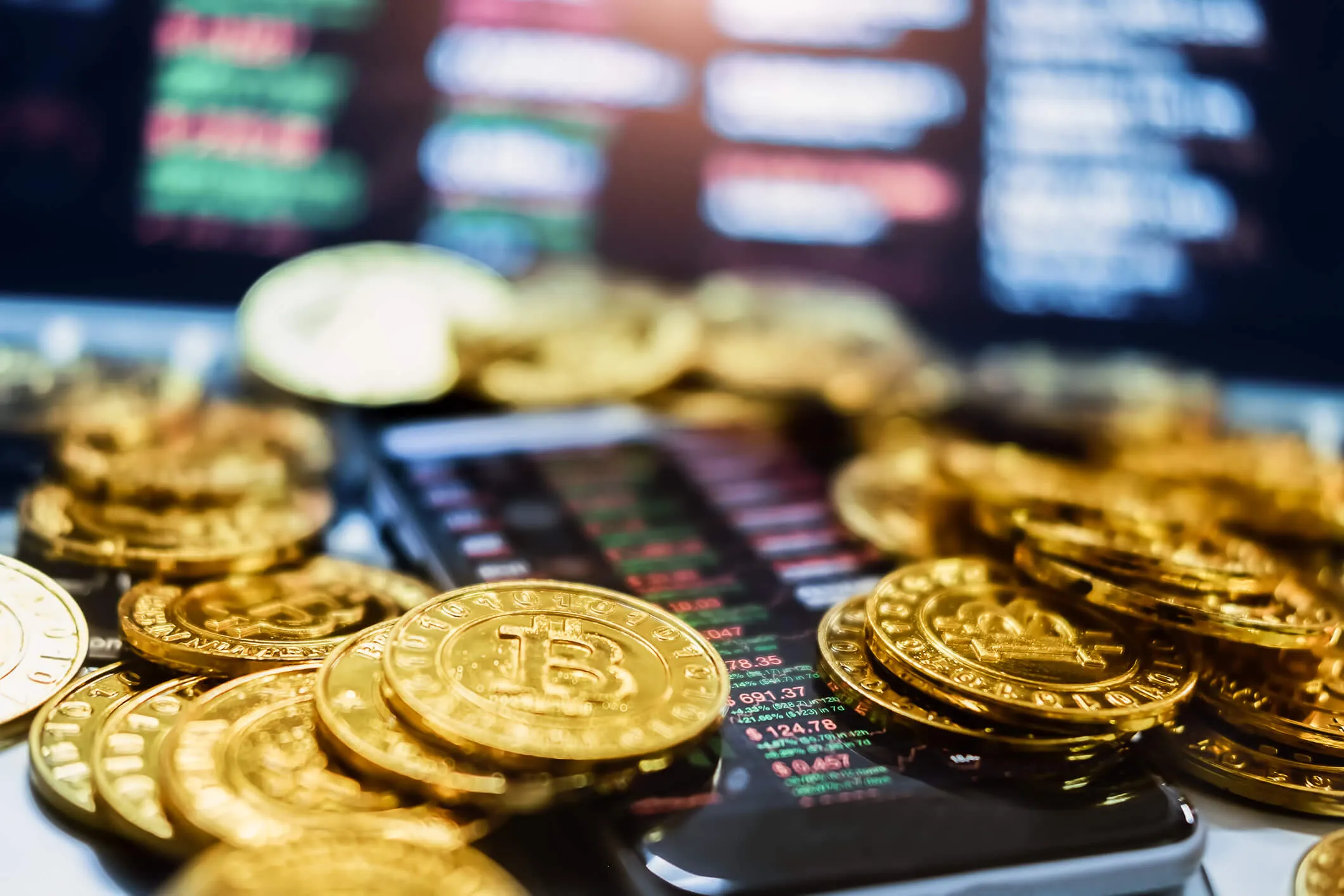 Close-up of golden Bitcoin coins scattered on a smartphone displaying cryptocurrency price charts with green and red numbers.