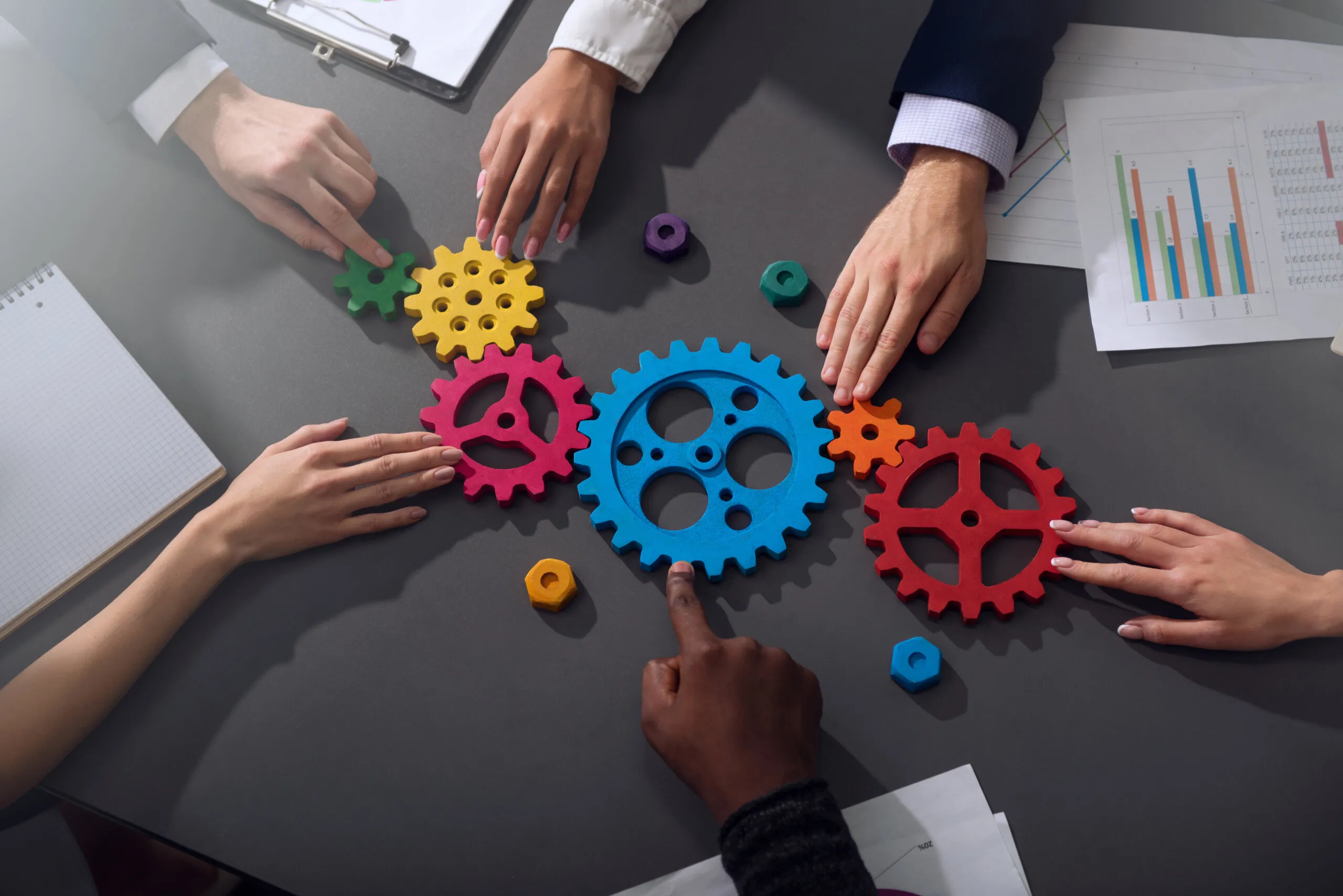 Five diverse hands collaboratively touching colorful wooden gears arranged on a table, symbolizing teamwork and collaboration, with papers and notebooks nearby.