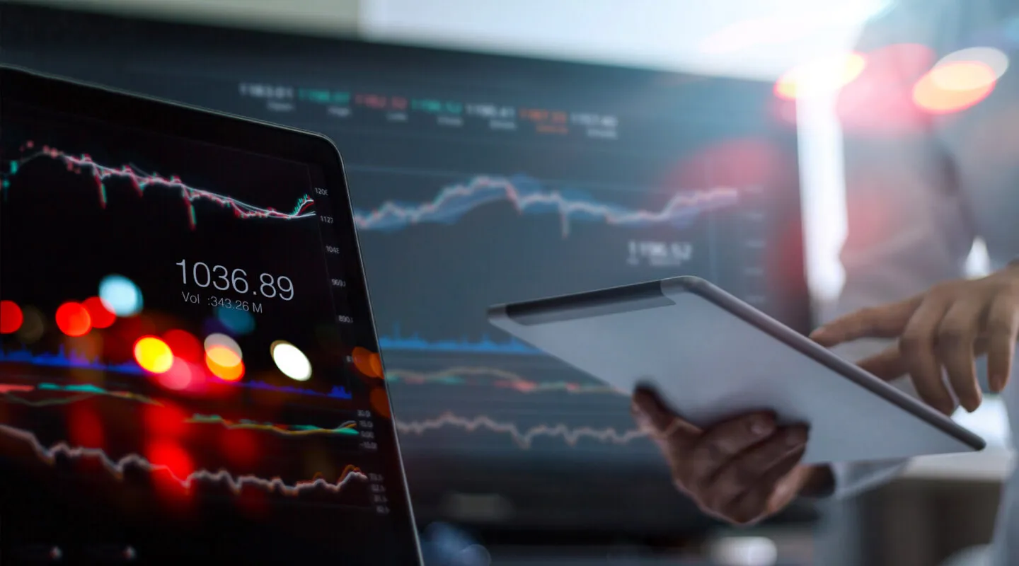 Person holding and interacting with a tablet in front of computer screens displaying financial charts and stock market data with numbers and graph lines.
