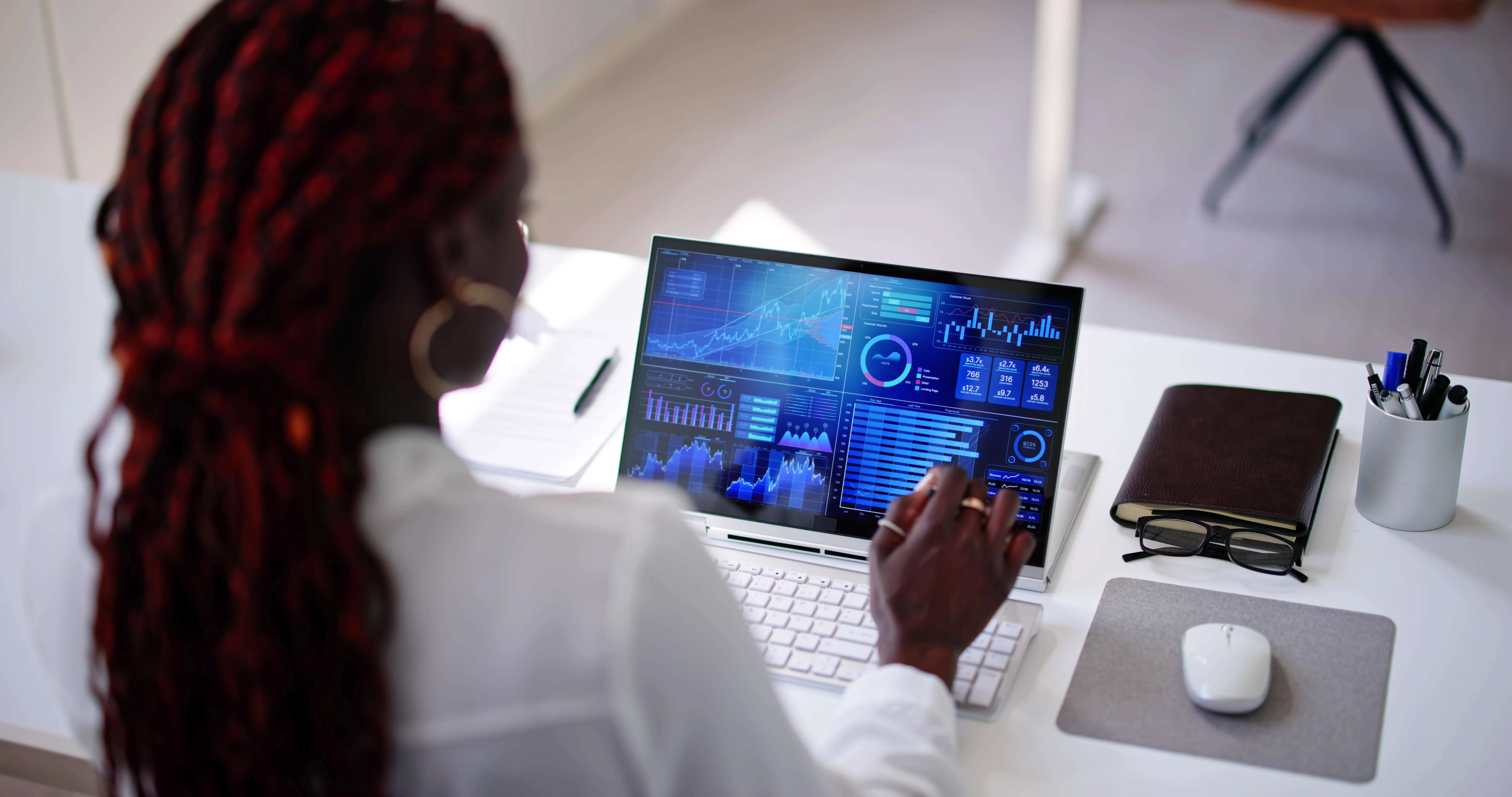 Person with braided red hair and gold hoop earrings sitting at a white desk, analyzing blue-themed data charts and graphs on a laptop screen, with a notebook, glasses, and a pen holder nearby.