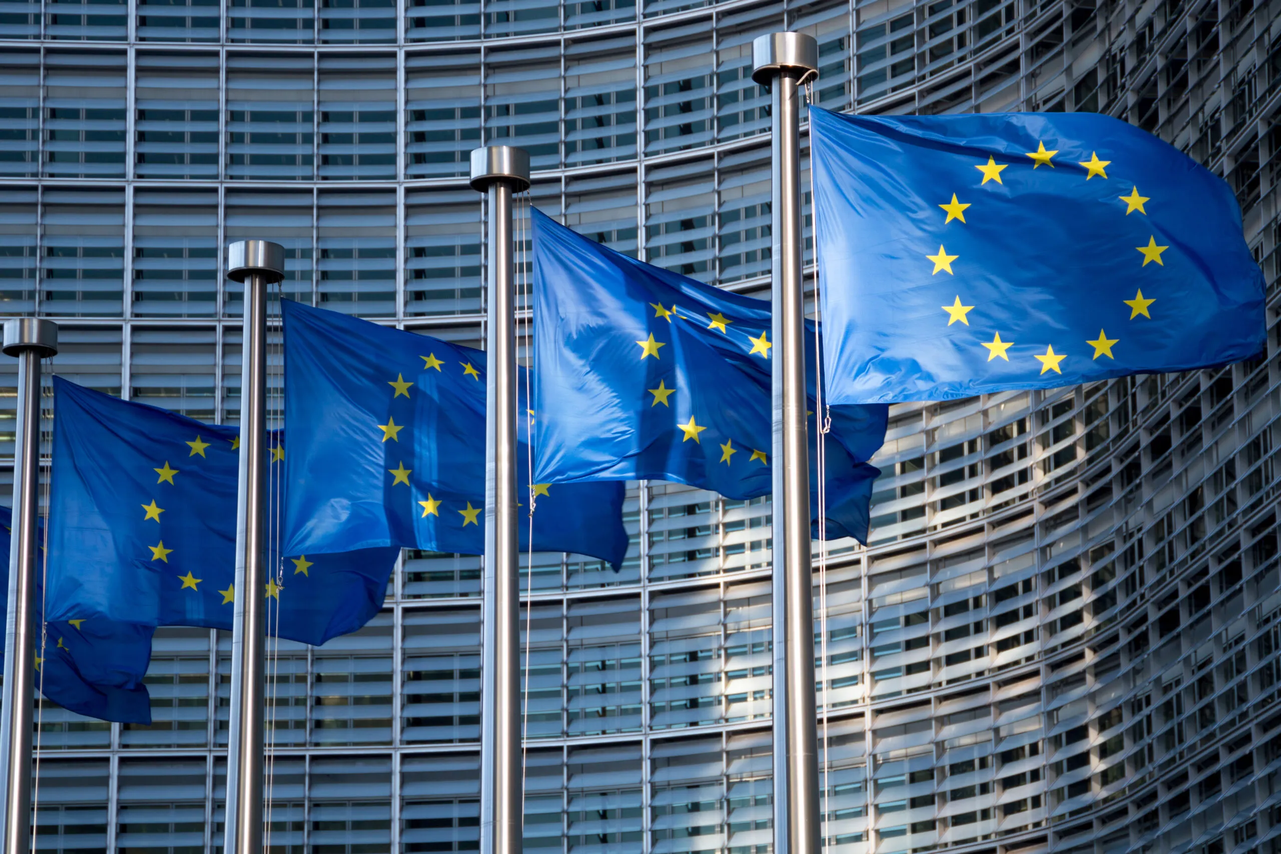 Several European Union flags waving on flagpoles in front of a modern glass building facade.