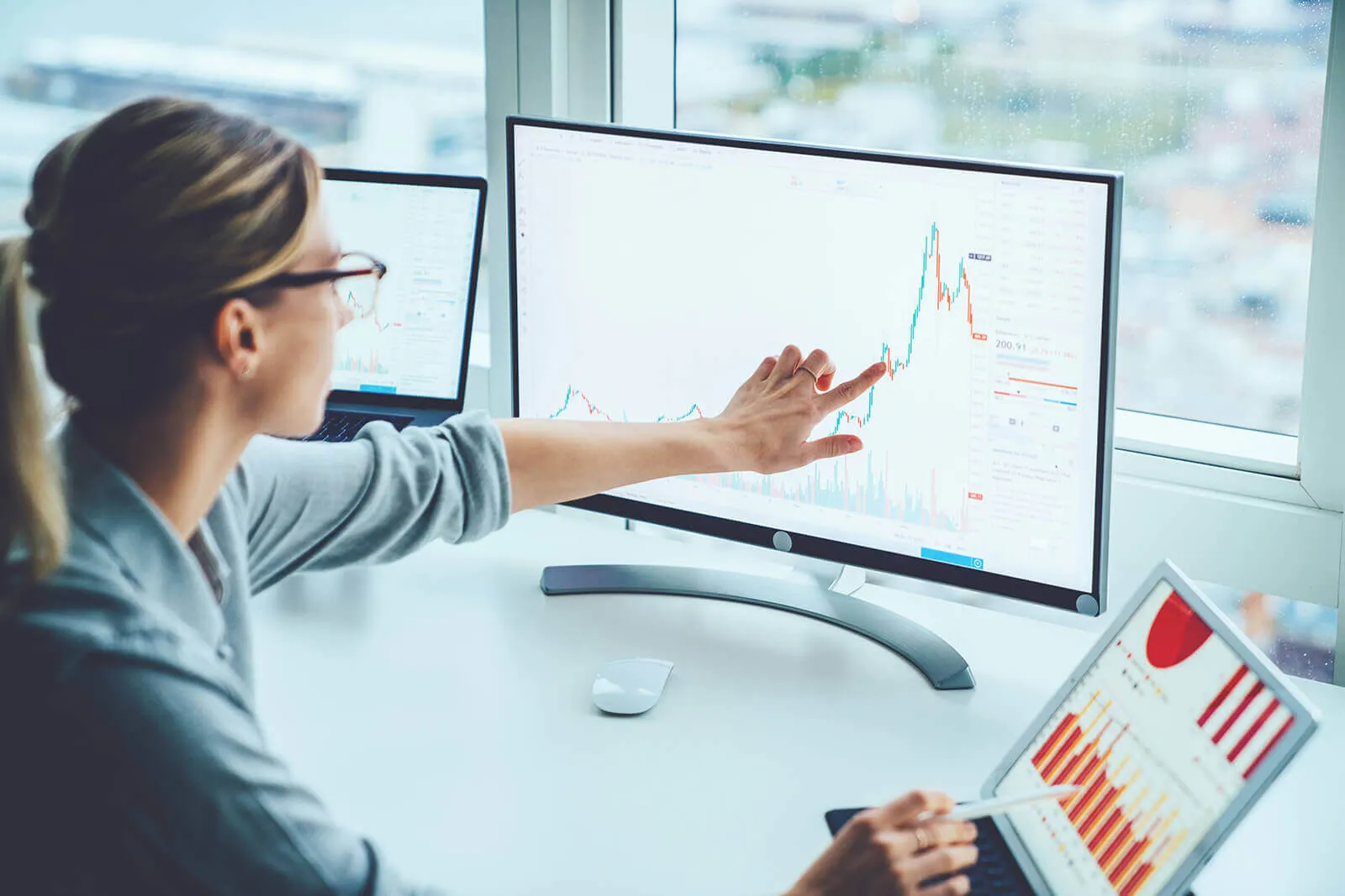 A woman wearing glasses analyzes financial charts on a large monitor, pointing at a rising graph line, with two additional screens showing data and bar charts around her in a bright office.
