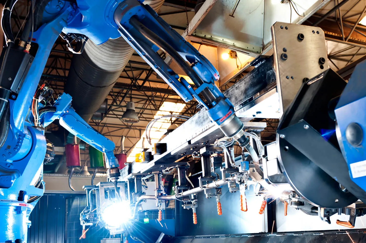 Close-up of blue industrial robotic arms welding metal pieces on an automated production line inside a factory with visible sparks and overhead lighting.