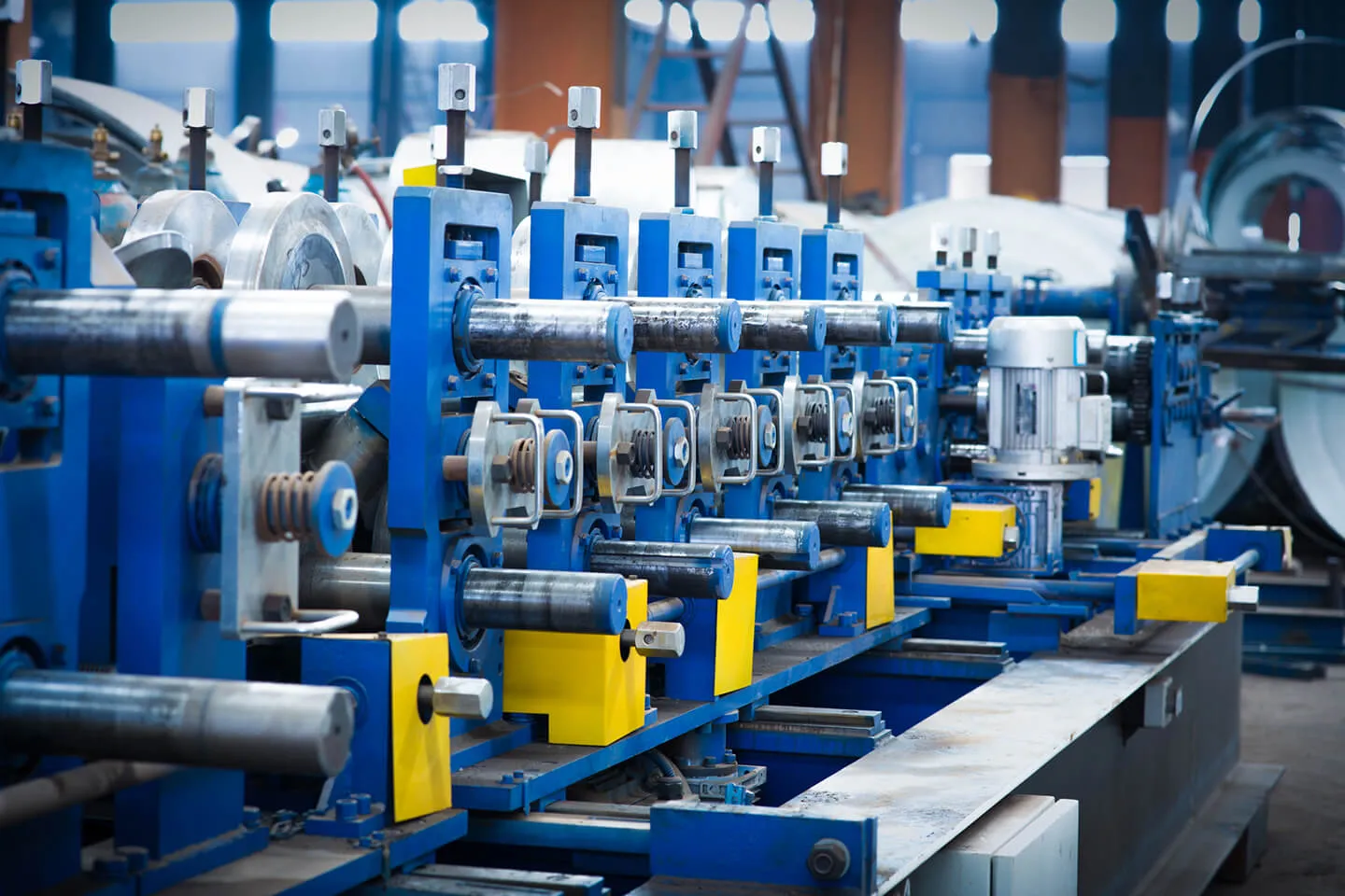 Close-up of a blue and yellow industrial machine in a factory with multiple cylindrical rollers and a motor on the right side, used for processing materials.