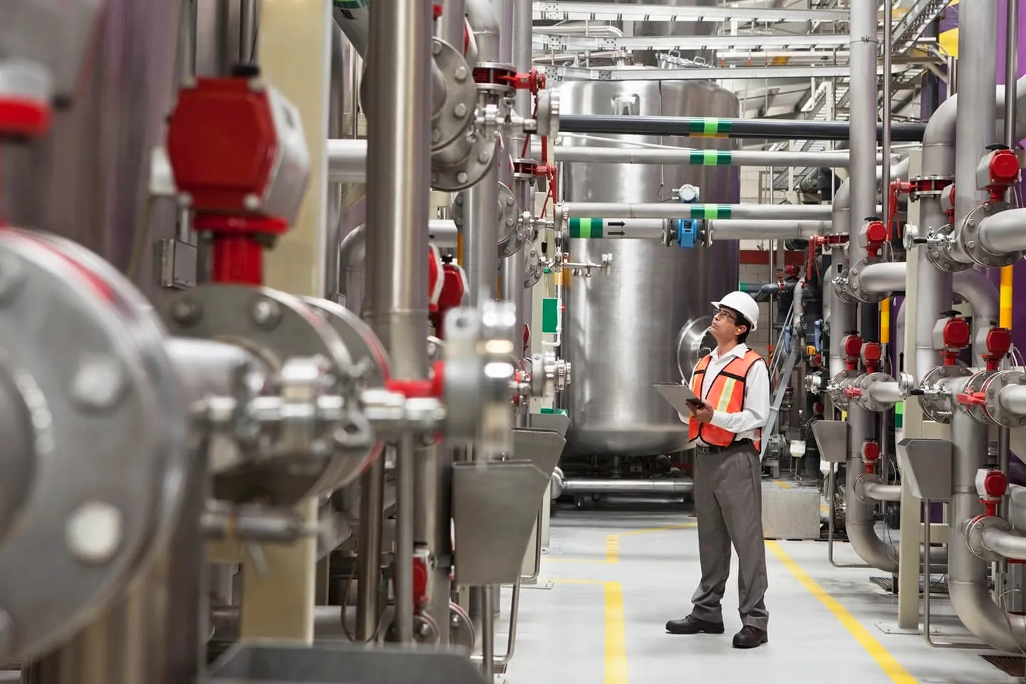 Engineer in safety vest and hard hat inspecting industrial pipes and tanks inside a facility, holding a tablet.