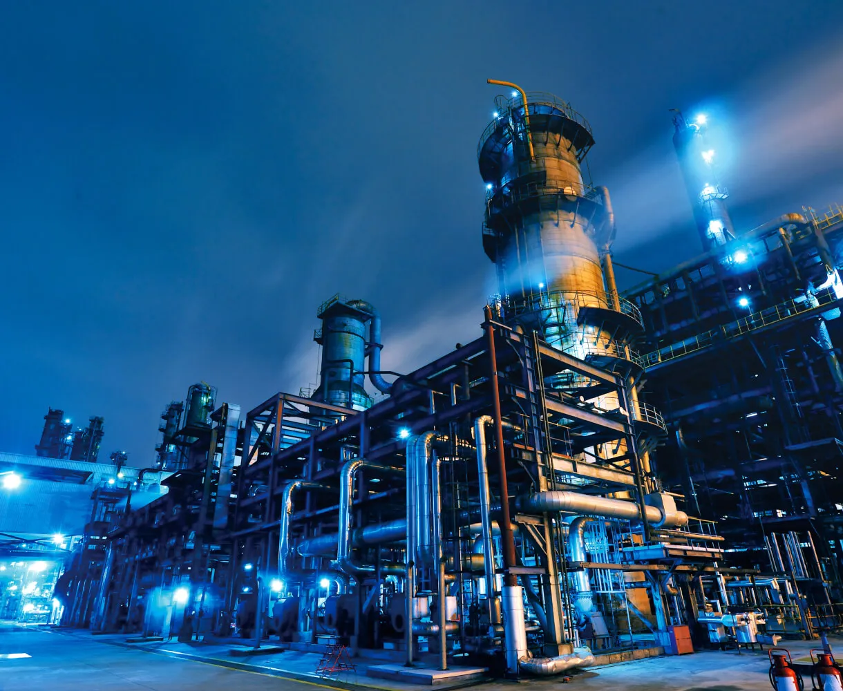 Industrial refinery or chemical plant at night with large towers, metal pipes, and bright blue lights illuminating the structure.