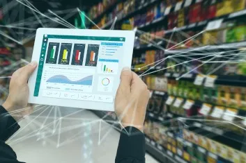 Person holding a tablet displaying various colorful charts and graphs in a grocery store aisle with blurred shelves of products in the background.